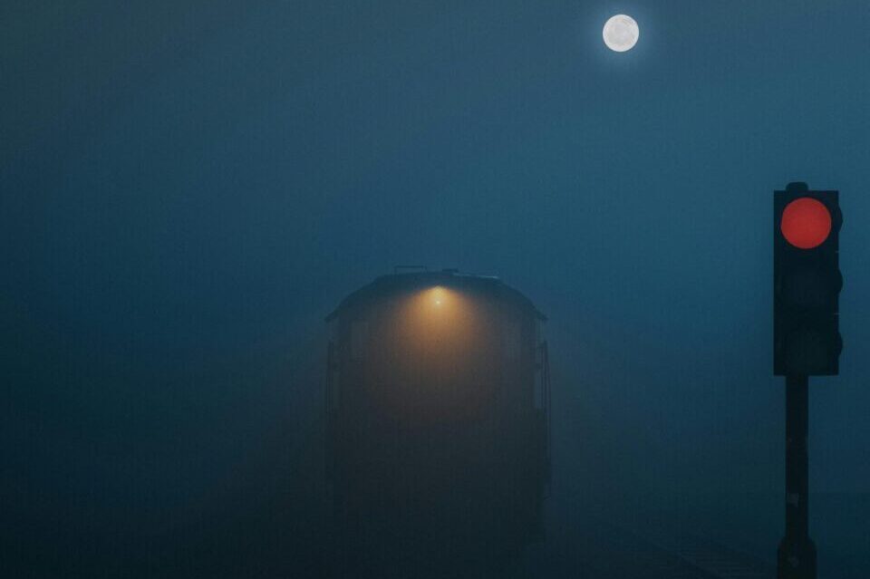 Atmospheric train scene in foggy night with full moon and red signal light, creating a mysterious mood.